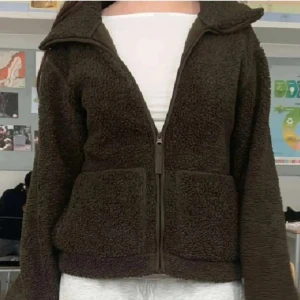 Grön teddyjacka med dragkedja - Mysig grön teddyjacka med stor krage och två rejäla fickor framtill. Jackan har en rak passform och stängs med dragkedja. Perfekt för kyliga dagar när du vill vara både varm och snygg. Materialet är fluffigt och ger en avslappnad vibe.