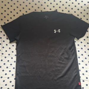 Svart Under Armour t-shirt S - Svart t-shirt från Under Armour i storlek S med klassisk logga på bröstet. Tillverkad i mjuk och lätt blandning av 70% bomull och 30% polyester, perfekt för träning. Enkel design med rund hals och korta ärmar, passar dig som gillar sportig och clean stil.