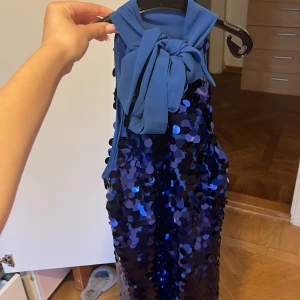 Blå paljettklänning med rosett - Säljer en glittrig blå kortklänning med stora paljetter och snygg rosett vid halsen. Klänningen har ärmlös design och en cool, lite loose passform. Perfekt för dig som vill sticka ut och glänsa med en unik look. Materialet är polyester och satin för extra skön känsla.