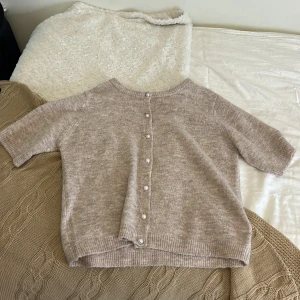 Beige kofta med pärlknappar från 157 - Supermjuk och mysig beige kofta från 157 med pärlknappar framtill. Koftan har kortare ärmar och är stickad i ett fluffigt material. Perfekt att slänga över ett linne för en chill look. Passar dig som gillar en lite loose fit.