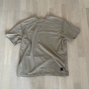 Beige basic t-shirt i bomull - Enkel och stilren beige t-shirt med korta ärmar. T-shirten har en klassisk passform och är tillverkad i mjuk bomull. Perfekt för en clean och avslappnad look. Liten svart etikett nertill på sidan ger en diskret detalj. Skriv vid frågor!🤝🏽