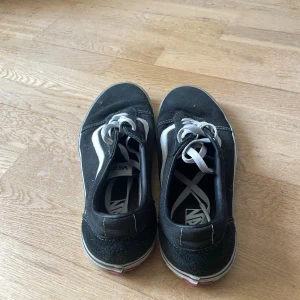 Svarta Vans Old Skool sneakers - Klassiska Vans Old Skool sneakers i svart med vit sidosnöre och vit sula. Skorna har snörning, rund tå och är tillverkade i en mix av mocka och canvas. Perfekta för en avslappnad streetstil och passar till det mesta i garderoben.