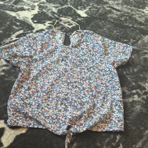  Blå topp med knytning  -  En blå blommig topp med en knytning i fram och ostretchning material som kan tillhöra en look med snygga Vibes 