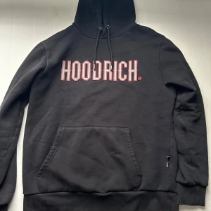 Svart HOODRICH sett - Svart hoodie från HOODRICH med stor vit och röd logga på bröstet. Tröjan har huva med snörning, magficka och långa ärmar. Materialet är mjuk bomull och passformen är normal. Perfekt för dig som gillar streetwear och vill ha en clean look. 