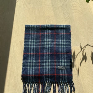 Blårutig halsduk från Burberry - Snygg halsduk från Burberry i klassiskt rutig design med marinblå, ljusblå och röda ränder. Gjord av 100% lammull, mjuk och varm med fransar i ändarna. Perfekt för att ge din outfit en cool och tidlös vibe.