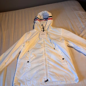 Vit vindjacka från Moncler med huva - Snygg vit vindjacka från Moncler med huva och dragsko. Jackan har hel dragkedja framtill, två fickor med knapp och Moncler-logga på insidan. Huvan har coola ränder i rött och svart för extra stilpoäng. Perfekt för dig som vill ha en clean och sportig look.