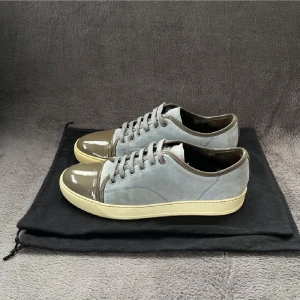 Grå mockasneakers med lackdetaljer - Snygga sneakers i grå mocka med glansig mörkgrå lack på tån och häl. Skorna har snörning och en beige platt sula som ger en clean look. Perfekta för dig som gillar stilrena men unika detaljer.