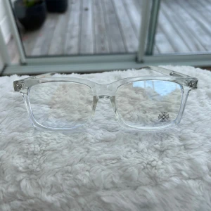 Chrome Hearts blåljus glasögon  - Säljer ett par stilrena glasögon från Chrome Hearts med transparent båge och rektangulär form. Bågarna har snygga silverdetaljer och små blommor på sidorna samt på glaset. Perfekt för dig som vill ha en unik och modern look.