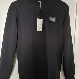 Svart sweatshirt från Valentino - Svart sweatshirt från Valentino med rund halsringning och diskret logopatch på bröstet. Tröjan har en klassisk passform och är tillverkad i mjuk bomull, perfekt för en clean och stilren look.