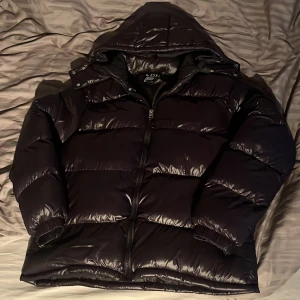 Svart pufferjacka från Brave Soul - Säljer en svart pufferjacka från Brave Soul i storlek M. Jackan har glansig finish, huva och dragkedja framtill. Perfekt för kalla dagar med sin rejäla vaddering och klassiska look. Passar dig som vill ha en stilren och varm jacka.