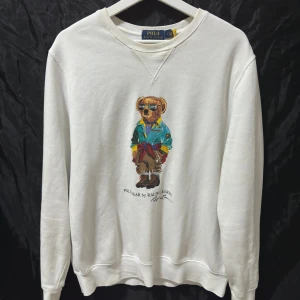 Vit sweatshirt Polo Ralph Lauren björn - Vit sweatshirt från Polo Ralph Lauren med ikoniskt björntryck på bröstet. Tröjan har rund hals, ribbade muddar och är långärmad. Tillverkad i mjuk bomull för en skön känsla. Perfekt för dig som gillar streetstyle och klassiska märken. Den är riktigt snygg men har tyvärr några få skavanker som man ser på sista bilden och näst sista. (Är likadant vid båda armarna) detta har inte hindrat mig från att använda den dock för det snyns knappt. Storlek M skick 8/10.