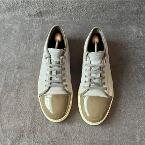 Grå sneakers med lackad tå - Snygga grå sneakers med ovandel i mocka och unik lackad tå i olivgrönt. Skorna har snörning och en ljus, platt sula som ger en clean look. Perfekta för dig som gillar stilrena detaljer och vill sticka ut lite extra.