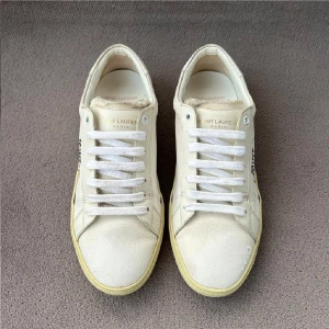 Saint Laurent vita sneakers i canvas - Snygga vita sneakers från Saint Laurent med handskriven logga på sidan. Skorna har klassisk låg profil, rund tå och vita snören. Ovandelen är i canvas med detaljer i skinn och sula i beige ton. Perfekta för en clean och avslappnad look.