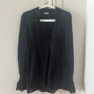 Svart långärmad blus från Hunkedory - Svart henley-blus från Hunkydory med v-ringning och broderad detalj framtill. Långärmad med volang vid ärmslut och lätt transparent material som ger en cool vibe. Perfekt för dig som gillar stilrena plagg med twist.