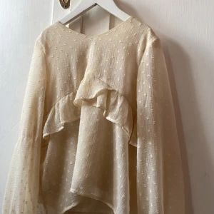 Beige prickig blus med volang - Säljer en superfin beige blus med diskreta prickar och volangdetaljer framtill. Blusen har lång ärm och är gjord i ett lätt, transparent tyg som ger en luftig känsla. Perfekt för dig som gillar romantiska och trendiga plagg med det lilla extra.