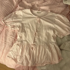 Ljusrosa blus med knyt från Gina Tricot - Supersöt ljusrosa blus från Gina Tricot med korta puffärmar och två knyt framtill. Blusen har en volangkant nedtill som ger en flowy känsla och är gjord i mjuk bomull. Perfekt för dig som gillar romantiska och feminina detaljer.