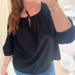 Svart blus med knyt i halsen - Säljer en svart blus med rundad hals och snygg knytning framtill. Blusen har trekvartsärmar med lätt puff och är gjord i ett mjukt, lätt material som faller fint. Perfekt för dig som gillar en enkel men stilren look.