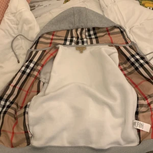 Grå Burberry hoodie med dragkedja - Snygg grå hoodie från Burberry i storlek M med klassiskt rutigt foder i huvan och insidan. Tröjan har dragkedja, snörning vid huvan och broderad Burberry-logga på bröstet. Perfekt för dig som gillar stilrena detaljer och ikoniskt mönster. Skulle kunna diskutera priset i sms