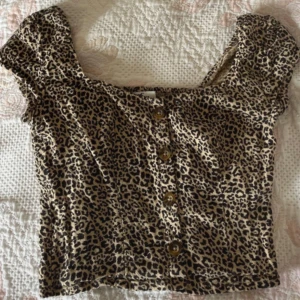 Leopardmönstrad topp från Hollister - Säljer en kortärmad leopardmönstrad  croptopp från Hollister med knappar framtill. Toppen har fyrkantig ringning, smockad rygg och är i mjuk bomullsmix. 