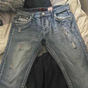 GS Premium blå jeans med slitningar - Säljer ett par blå jeans från GS Premium med coola slitningar och detaljerade bakfickor med kontrastsömmar. Jeansen har en klassisk femficksdesign, silverfärgade knappar och en snygg tvättad look. Perfekta för dig som gillar streetstyle och vill sticka ut.                               Köpt i USA för 220 dollar 