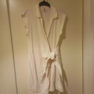 Vit omlottklänning Tommy Hilfiger - Vit kort omlottklänning från Tommy Hilfiger i mjukt bomullstyg. Klänningen har knyt i midjan, krage och är ärmlös med lätt volangkant. Klassisk och stilren design med diskret logga på ärmen. Perfekt för en fräsch och avslappnad look.