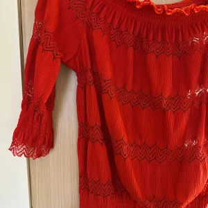 Röd offshoulder blus från River Island strl: 8 - Superfin röd offshoulder blus från River Island med smockad resår upptill och vid ärmslut. Blusen har långa ärmar, broderade spetsdetaljer och vågiga kanter. Materialet är lätt och luftigt, perfekt för att sticka ut med färg och mönster.