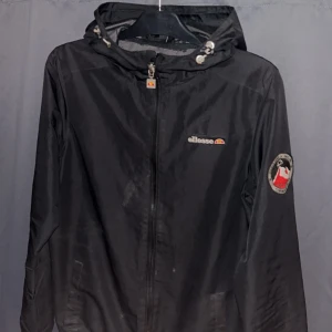 Svart vindjacka från Ellesse - Svart vindjacka från Ellesse med huva och dragkedja framtill. Jackan har logga på bröstet och ett märke på ena ärmen. Justerbar huva med snörning och praktiska fickor. Perfekt för blåsiga dagar.