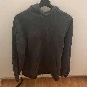 Mörkgrå fleecehoodie från Adidas - Mysig mörkgrå hoodie från Adidas i fleece med broderad logga på bröstet. Tröjan har huva med snörning, känguruficka och långa ärmar. Perfekt för kyliga dagar när du vill vara både bekväm och stilren. Knappt använd