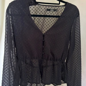 Svart prickig blus från Bik Bok XS - Säljer en svart transparent blus från Bik Bok i storlek XS. Blusen har små prickar i tyget, v-ringning, knappar framtill och volang nedtill. Långa ärmar och lätt figursydd passform. Tillverkad i 100% polyester.