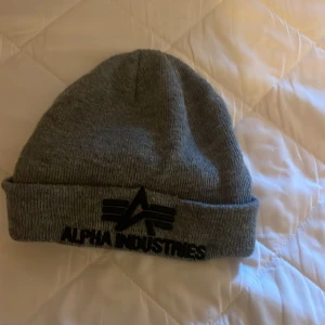 Grå mössa Alpha Industries - Snygg grå mössa från Alpha Industries med svart logotyp broderad framtill och liten etikett på sidan. Klassisk ribbstickad design med uppvikt kant, perfekt för kalla dagar. Passar dig som gillar streetwear och enkel stil.