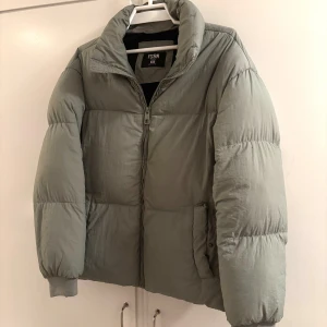 Grå pufferjacka från FSBN  - Säljer en grå pufferjacka från FSBN i storlek XS. Jackan har hög krage, dragkedja framtill och två fickor med knappstängning. Perfekt för kalla dagar med sin vadderade och oversized look. Passar S/M. 