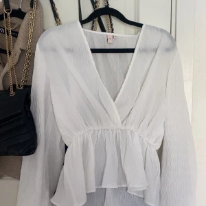 Vit omlottblus från Nelly, stl 38 - Supersöt vit omlottblus från Nelly i storlek 38. Blusen har v-ringning, långa ärmar och volang nedtill. Tunt, lätt och strukturerat tyg som ger en luftig känsla. Perfekt att styla med jeans eller kjol för en fräsch look.