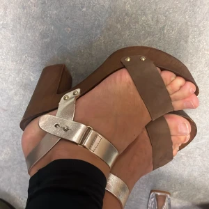 Replay Bruna sandaler med klack och metallicrem - Snygga sandaler med träfärgad klack och plattformsula. Bred rem i brunt skinn över tårna och metallicfärgad rem runt ankeln med spänne. Tillverkade i Italien, perfekt för dig som gillar en retrovibe och vill sticka ut med metallicdetaljer.