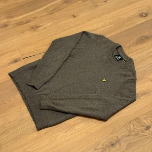 Grön tröja från Lyle & Scott - Snygg grön stickad tröja från Lyle & Scott med rund halsringning och klassisk gul logga på bröstet. Tröjan har ribbade muddar vid ärmslut och nederkant, perfekt för lager-på-lager under kyliga dagar.