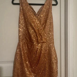 Glittrig guldig kortklänning - Säljer en superglittrig guldig kortklänning med omlott framtill och djup v-ringning. Klänningen är ärmlös och har snyggt korsade axelband i ryggen. Perfekt för dig som vill sticka ut och glänsa på nästa fest!
