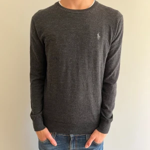 Mörkgrå merinoullströja Polo Ralph Lauren - Snygg mörkgrå tröja från Polo Ralph Lauren i tvättbar merinoull. Slim fit-modell med rund halsringning och diskret broderad logga på bröstet. Perfekt för dig som gillar stilrena och klassiska plagg med en modern passform.