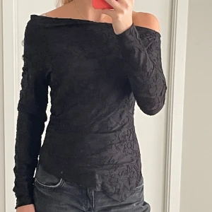 Svart offshoulder topp med struktur - Säljer en svart långärmad topp med offshoulder-modell och snyggt strukturerat tyg. Toppen har en tight passform och är perfekt för dig som gillar en stilren men ändå unik look. Materialet har en lätt mönstrad känsla som ger extra edge.