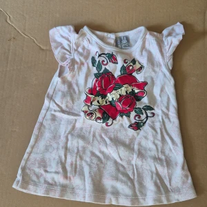 Vit topp med ros-tryck från Six Bunnies för barn - Supersöt vit topp från Six Bunnies med coolt tryck av röda rosor och banderoll framtill. Toppen har små volangärmar och diskret rosa mönster över hela tyget. Tillverkad i 100% bomull, perfekt för en unik och lekfull stil.