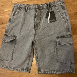 Grå cargoshorts från Brave Soul - Säljer ett par grå cargoshorts från Brave Soul med klassisk loose passform. Shortsen har stora sidofickor med lock, två bakfickor och bälteshällor. Materialet är jeans och färgen är en snygg ljusgrå ton som passar till det mesta.