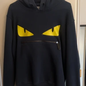Svart Fendi hoodie med gula ögon - Cool svart hoodie från Fendi med ikoniska gula ögon framtill och en dragkedja under ögonen. Tröjan har huva och långa ärmar, perfekt för dig som vill sticka ut. Mjuk och skön bomullskänsla, passar till streetwear-stilen.