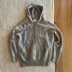 Davida Kashmir hoodie  - Mjuk och lyxig grå hoodie i kashmir från Davida. Tröjan har huva, dragsko och fickor framtill. Perfekt för en avslappnad och stilren look. Klassisk passform och ribbade muddar vid ärmslut och nederkant. Storlek är M men den är tvättad så storlek blir XS. Sydd vid armhåla men syns ej.