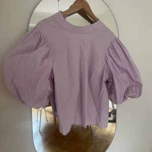 Ljuslila blus med puffärmar - Supersöt ljuslila blus med stora puffärmar och hög hals. Blusen har en knapp i nacken och är tillverkad i ett lätt bomullstyg. Perfekt för dig som vill sticka ut med en trendig och feminin look.