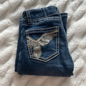 Blå jeans med broderad ficka Gina Tricot - Snygga blå jeans från Gina Tricot med låg midja och kontrastsömmar. Bakfickan har en broderad detalj i silver och guld med paljetter som ger extra edge. Klassisk femficksmodell i jeansmaterial, perfekt till en cool vardagslook.