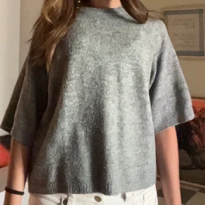 Grå stickad tröja med kort ärm - Snygg grå stickad tröja med korta, vida ärmar och rund hals. Perfekt för dig som gillar en chill och enkel stil. Tröjan har en loose passform och är superbekväm att bära till både jeans och kjol. Ribbad kant nedtill ger en extra touch.