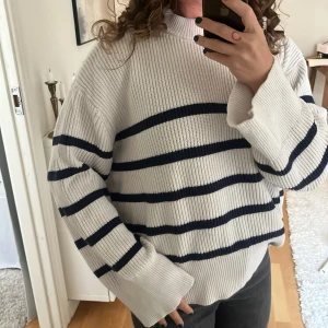 Randig stickad tröja från NA-KD - Supermysig stickad tröja från NA-KD i vitt med mörkblåa horisontella ränder. Tröjan har en högre ribbad krage, lång ärm och relaxed fit. Perfekt för lager-på-lager och chill dagar. Snygg till både jeans och kjol.