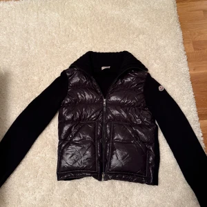 Svart dunväst från Moncler med stickade ärmar - Säljer en svart cardiganfrån Moncler med glansig quiltad framsida och två stora fickor, modell heter “padded Wool Cardigan” i Storlek S. Ärmar och rygg är stickade i mjukt material och kragen är ribbad. Klassisk Moncler-logga på ena ärmen. Köpt från Monclers hemsida. Inga tecken på användning, endast använt ett fåtal gånger.