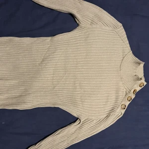 Beige ribbad tröja med knappar - Snygg beige ribbad tröja från H&M i storlek XS. Tröjan har lång ärm, hög hals och fyra dekorativa knappar på ena axeln. Den är figurnära och har en stilren look som passar perfekt till jeans eller kjol. Har använt kanske två gånger.