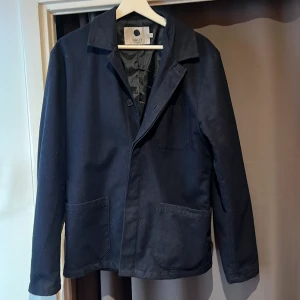 Mörkblå kavaj från NN07 - Snygg mörkblå kavaj från NN07 med klassisk krage och tre fickor framtill. Jackan har en stilren look och är tillverkad i ett mjukt bomullsmaterial. Perfekt för dig som gillar enkel och clean stil. Passar dig som vill ha en jacka med normal passform och lång ärm.