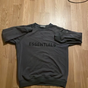 Svart Essentials Fear of God sweatshirt - Säljer en svart sweatshirt från Essentials Fear of God med tryck på bröstet och ärmen. Tröjan har rund hals, ribbade muddar och en loose passform. Perfekt för dig som gillar streetwear och vill ha en stilren look. Storlek L. Har använt många gånger men bra skick ändå!