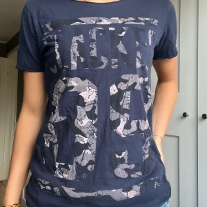 Blå t-shirt med camotryck  - Mörkblå t-shirt från 157 DOG med coolt camo-inspirerat tryck och stora bokstäver framtill. Klassisk passform med rund hals och korta ärmar. Tillverkad i mjuk bomull som är skön mot huden. Perfekt för en avslappnad streetstyle.